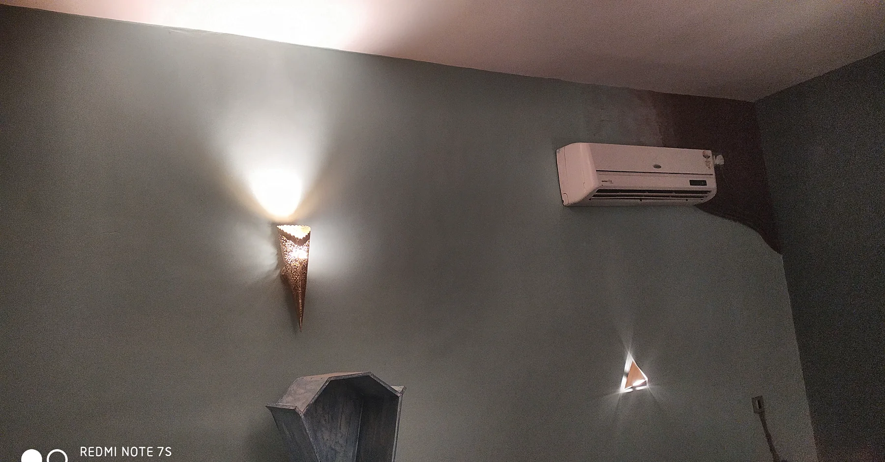 A wall lamp throws a cone of light up the wall, with a triangle light low beside a small wooden niche.